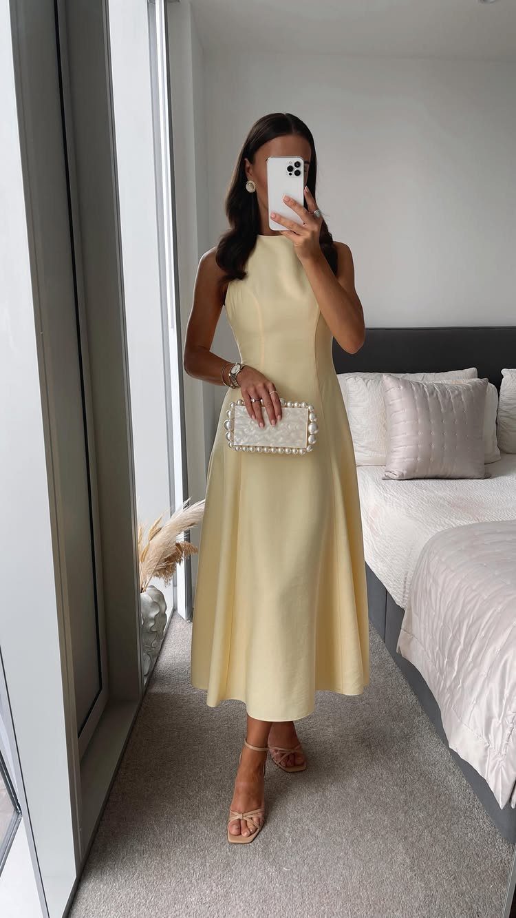 Woman in a pale yellow sleeveless midi dress holding a white beaded clutch in a bedroom.