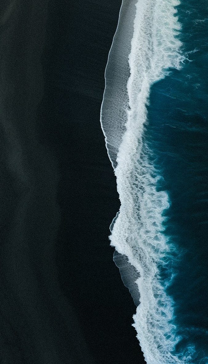 Black sand beach meeting deep blue ocean waves with white sea foam from an aerial view.