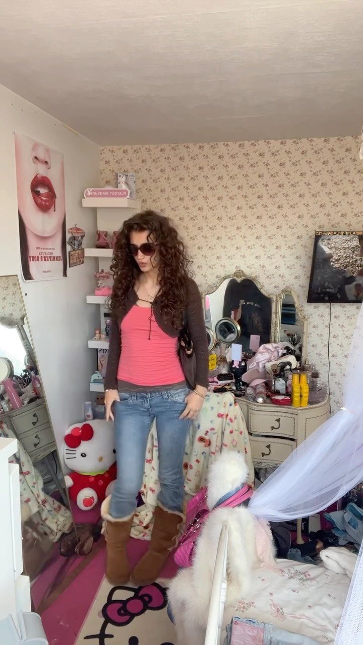 A woman posing in a bedroom wearing a pink tank top, low-rise jeans, and fuzzy boots.