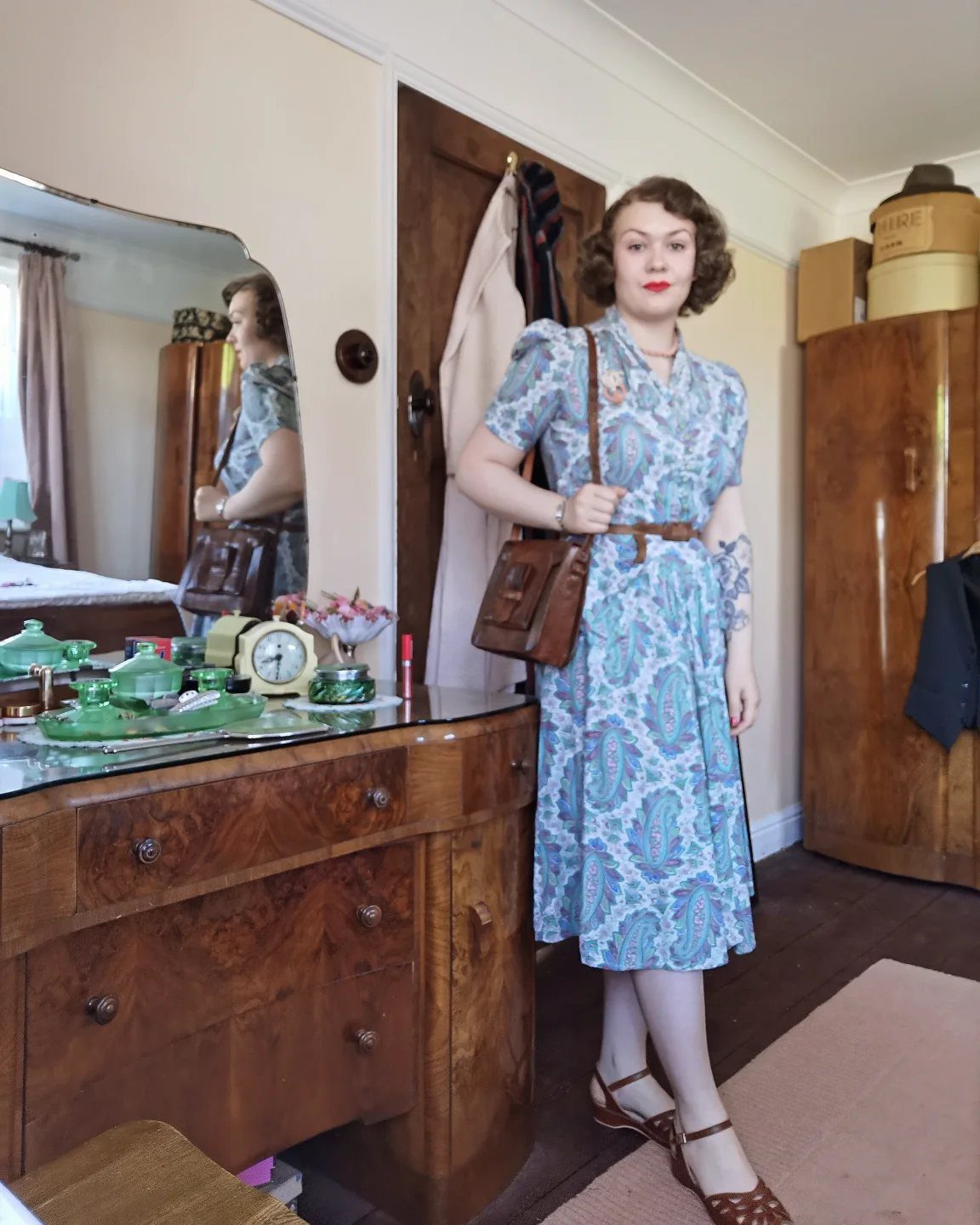 A woman posing in a blue patterned 1940s style dress and brown accessories.