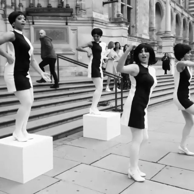 Women in black and white shift dresses dancing on white pedestals outdoors.