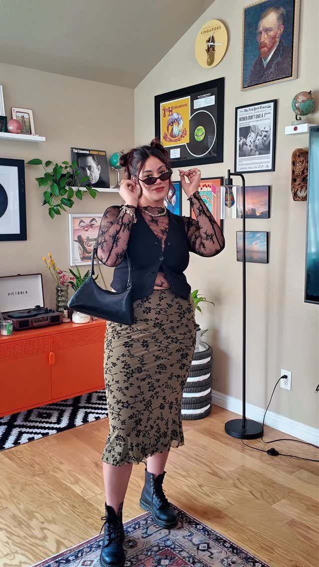 A woman wearing a black lace top, vest, floral skirt, and combat boots posing indoors.