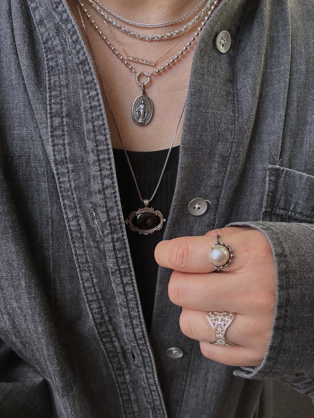 Layered silver necklaces with various pendants worn over a grey denim shirt and black top.