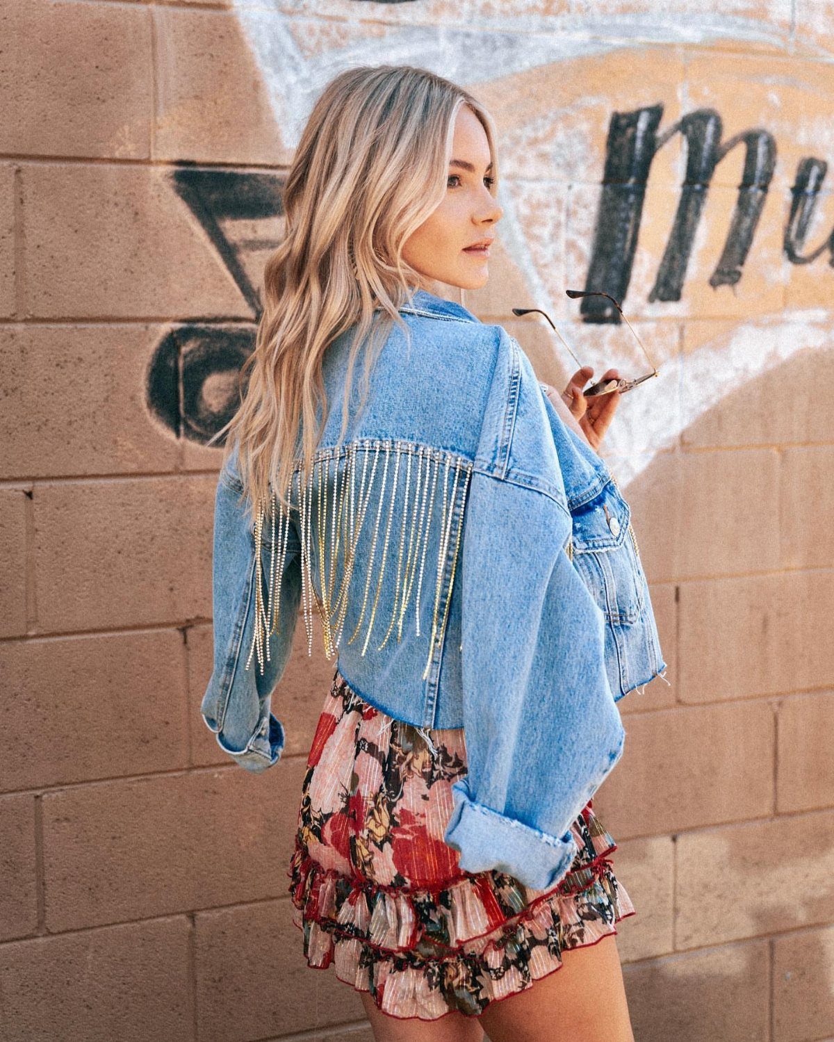 Blonde woman wearing a light wash denim jacket with long gold fringe detailing on the back.