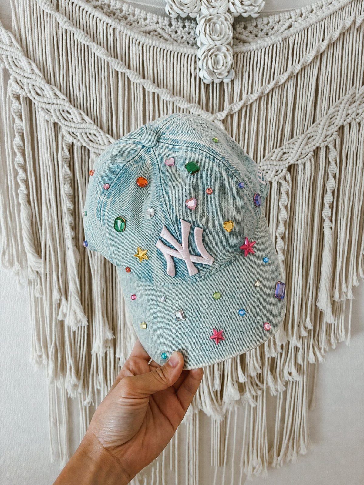 A light blue denim baseball cap embellished with colorful rhinestones held against a macrame background.