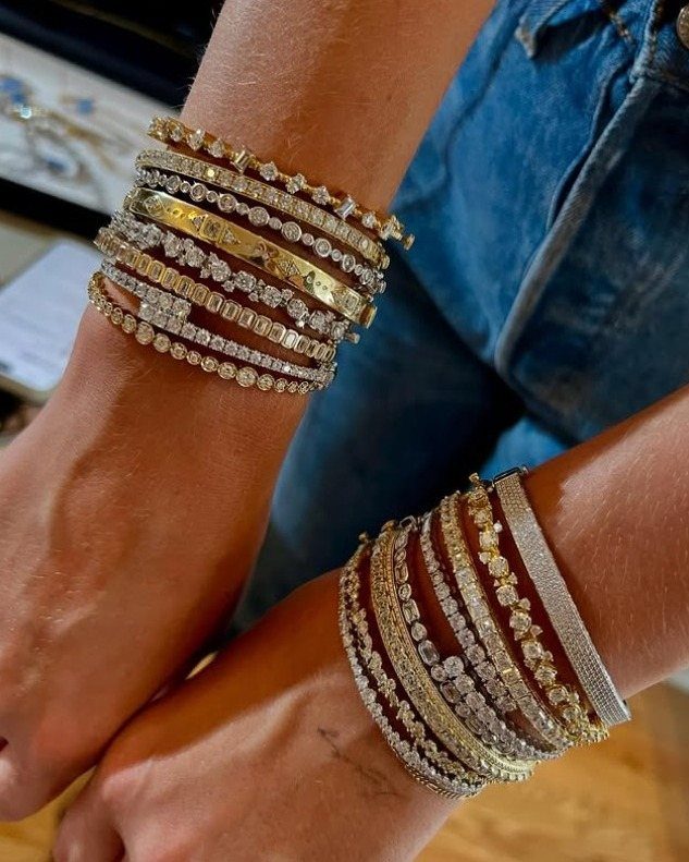 Multiple stacked gold and silver rhinestone bracelets worn on two wrists against blue denim.