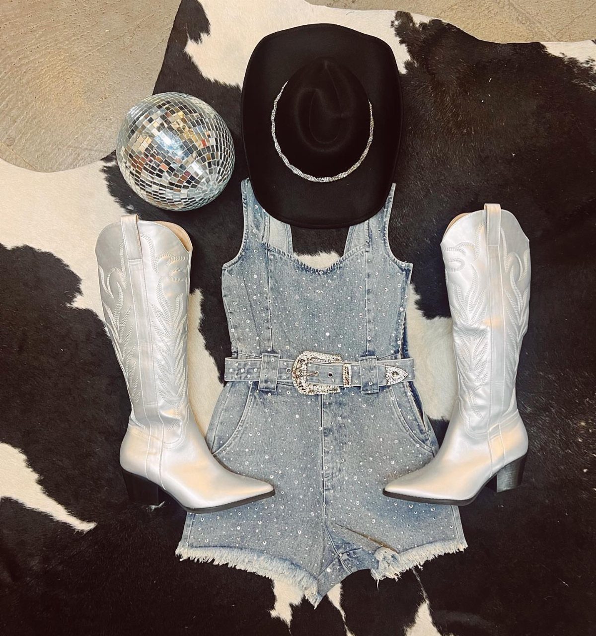 Denim romper with rhinestones, silver cowboy boots, and a black hat on cowhide.