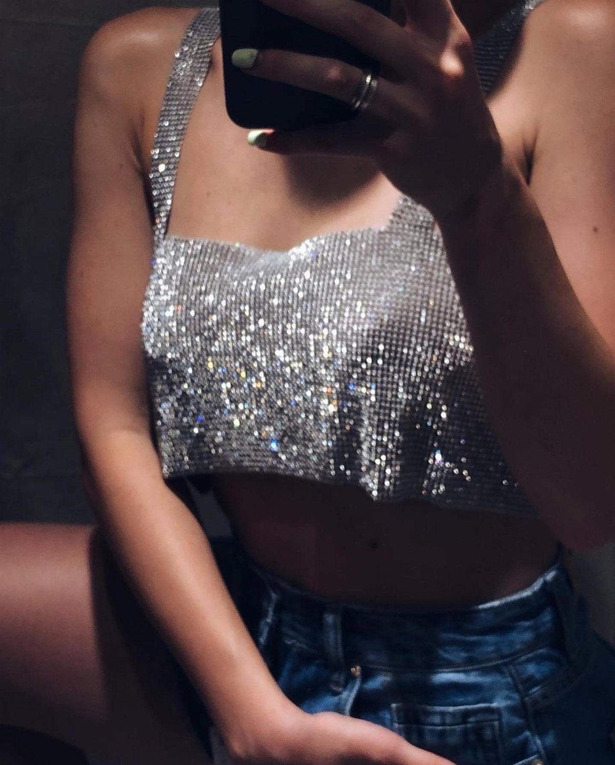 A person wearing a sparkling silver sequined camisole and blue denim shorts taking a selfie.