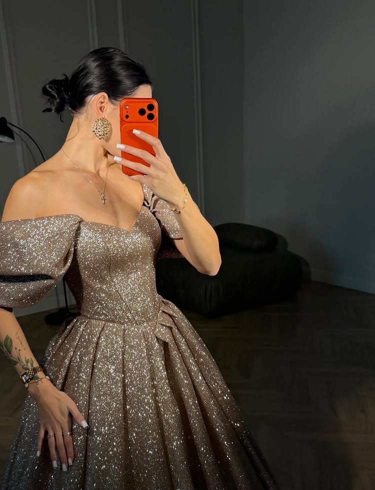 A woman posing in a shimmering champagne-colored glitter gown with off-the-shoulder sleeves.