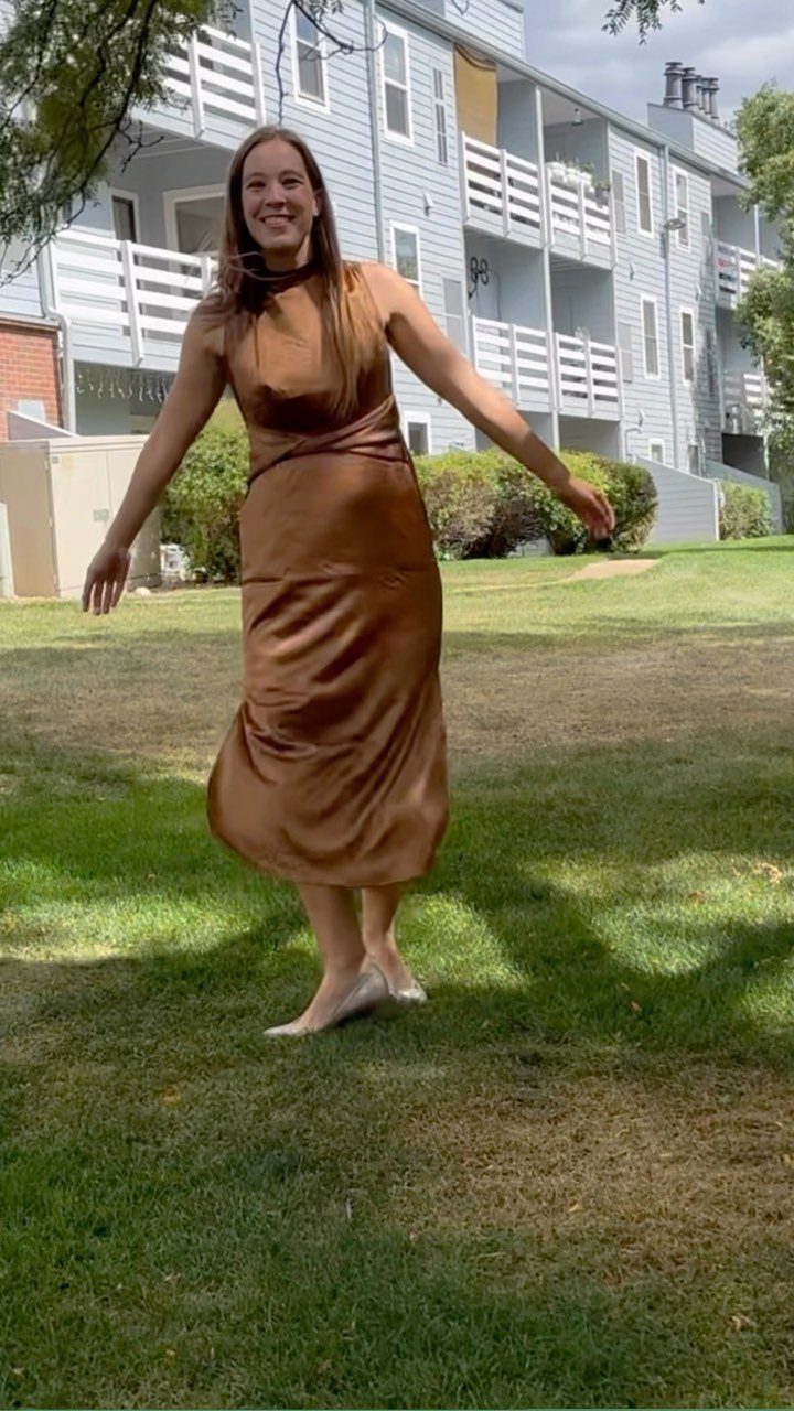 A woman wearing a brown satin midi dress standing on a green lawn outdoors.