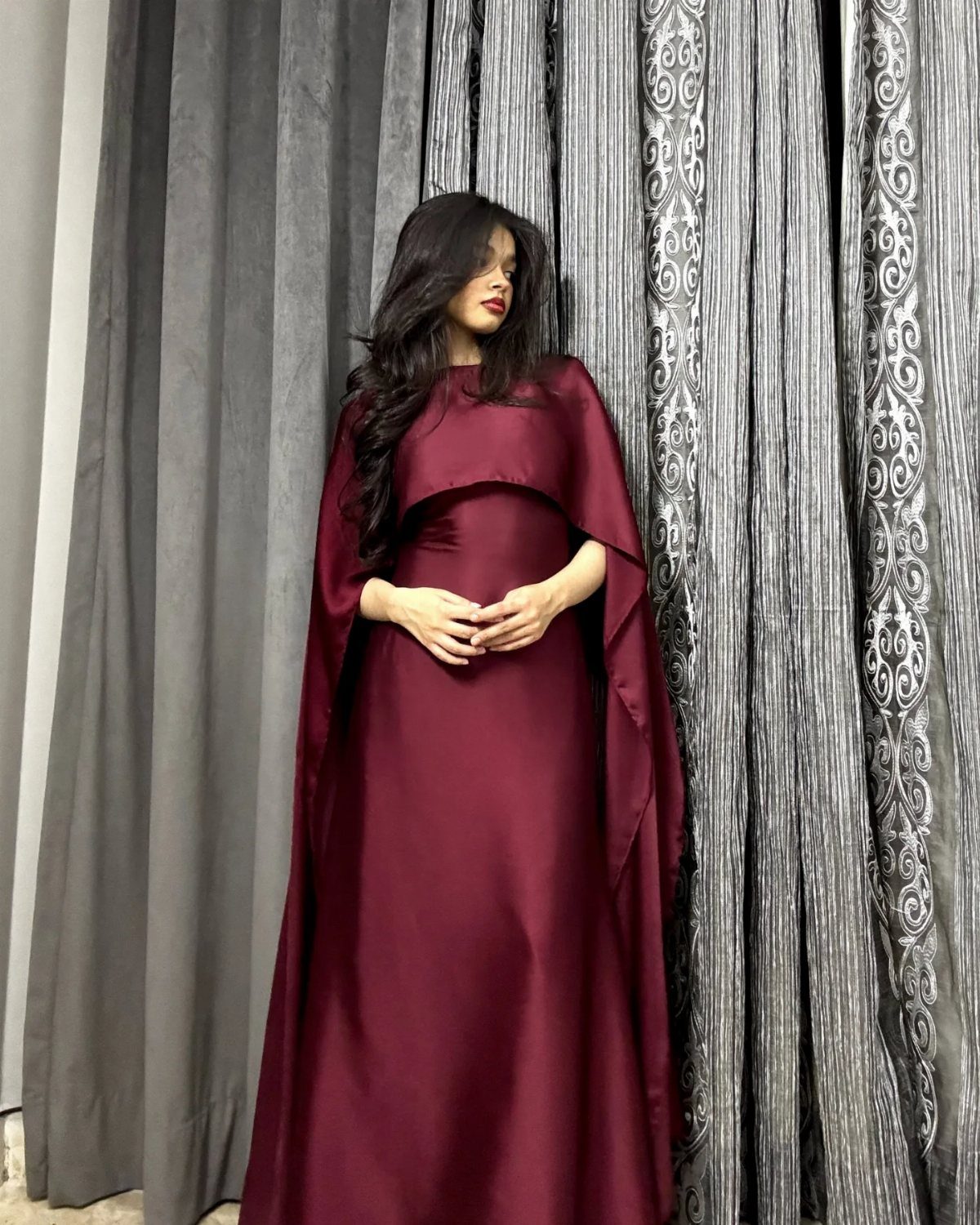 Woman in a long maroon satin cape dress standing against grey patterned curtains.