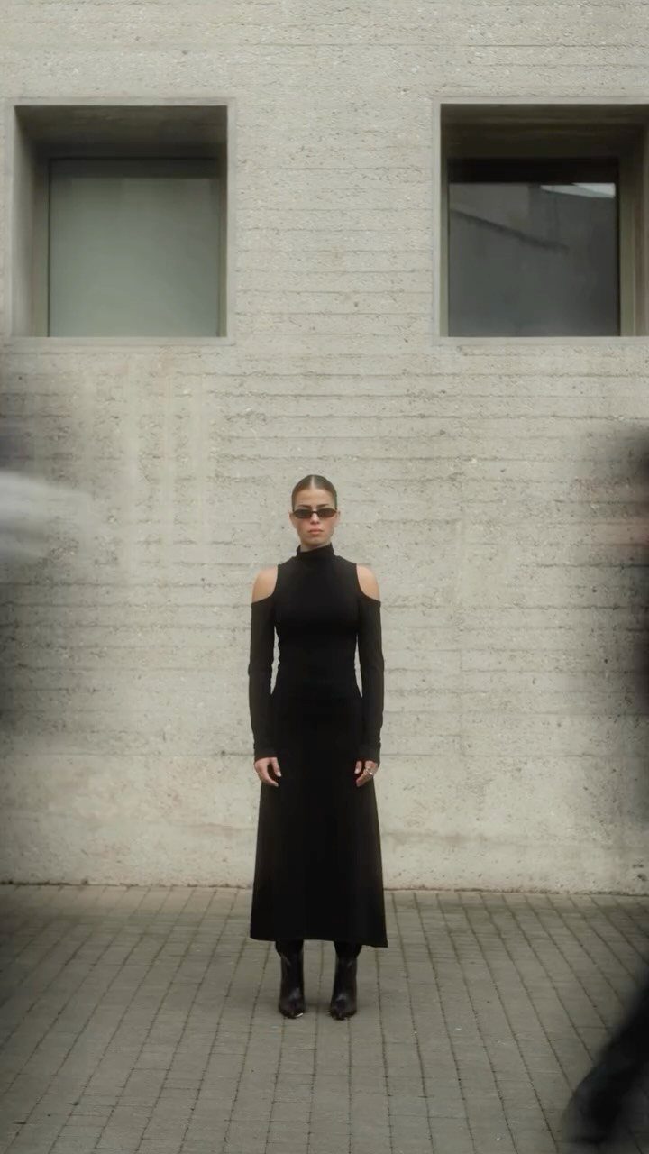 Woman in a black long-sleeved dress with shoulder cut-outs standing against a concrete wall.