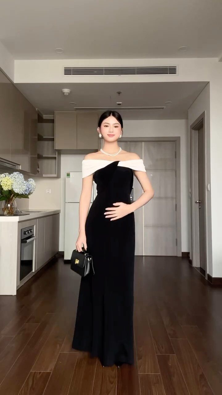 A woman posing in a long black off-the-shoulder evening dress inside a modern apartment.
