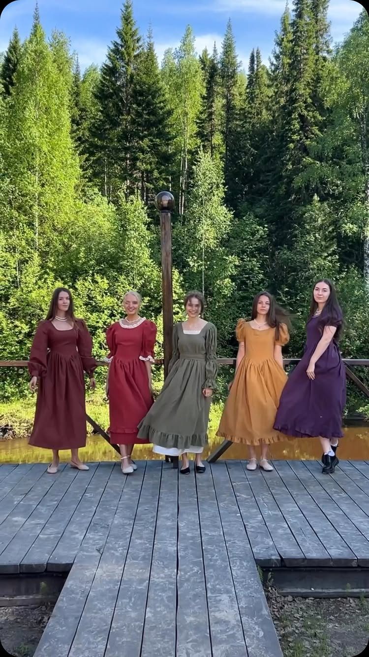 Five women standing on a wooden deck wearing colorful, loose-fitting peasant dresses in a forest.
