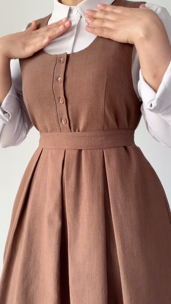 A person wearing a brown button-down pinafore dress over a white collared shirt.
