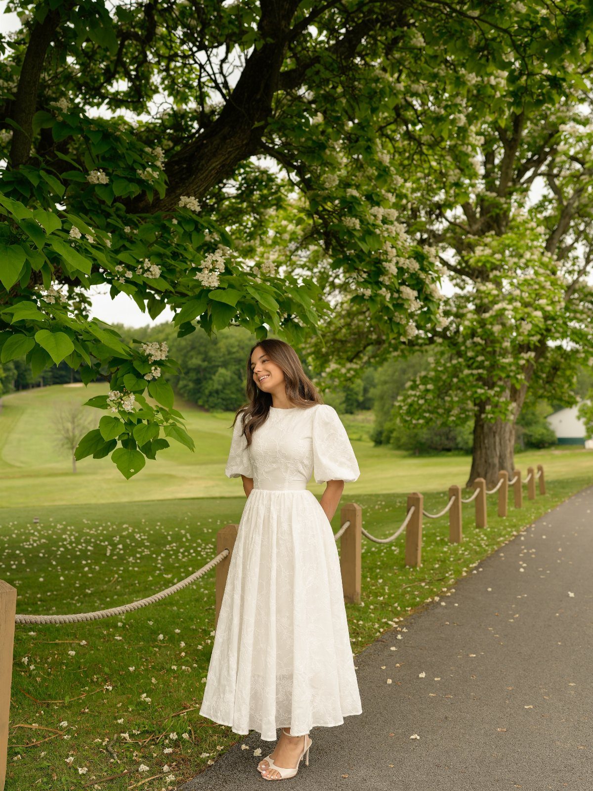 A woman in a white puff-sleeved midi dress standing on a path under a tree.