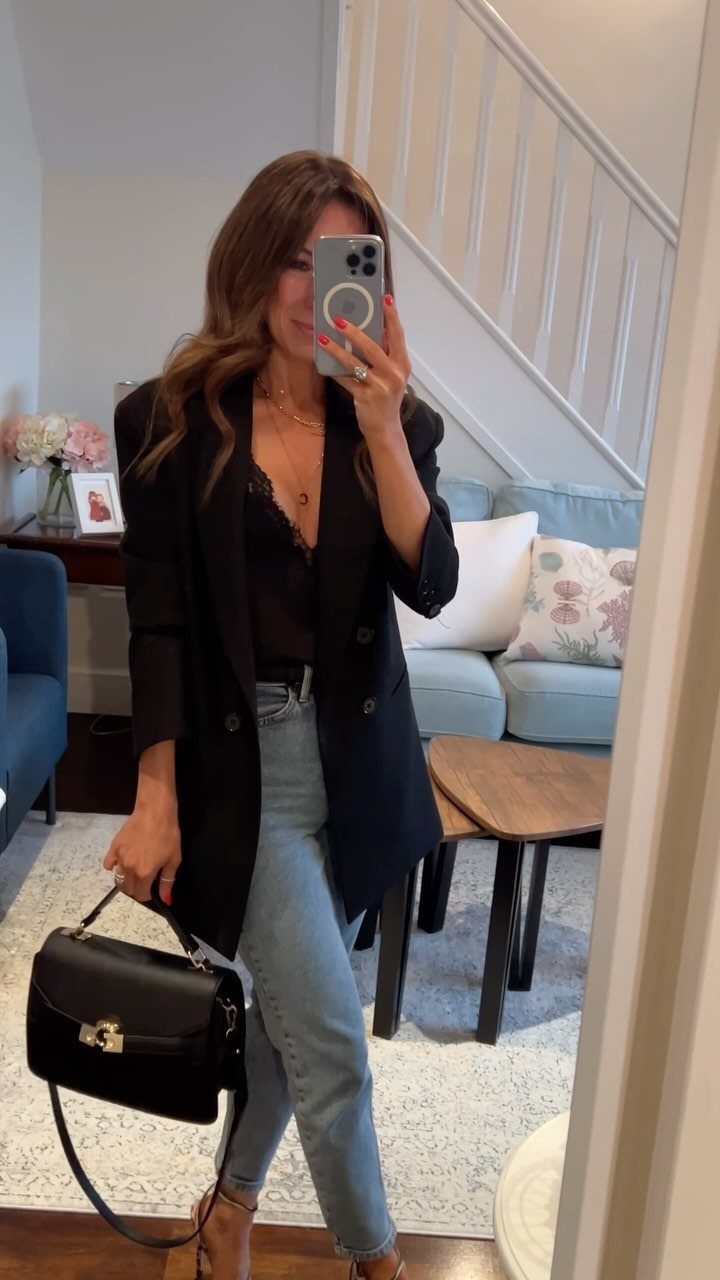 Woman in a black blazer, black lace top, and light blue jeans holding a black handbag.