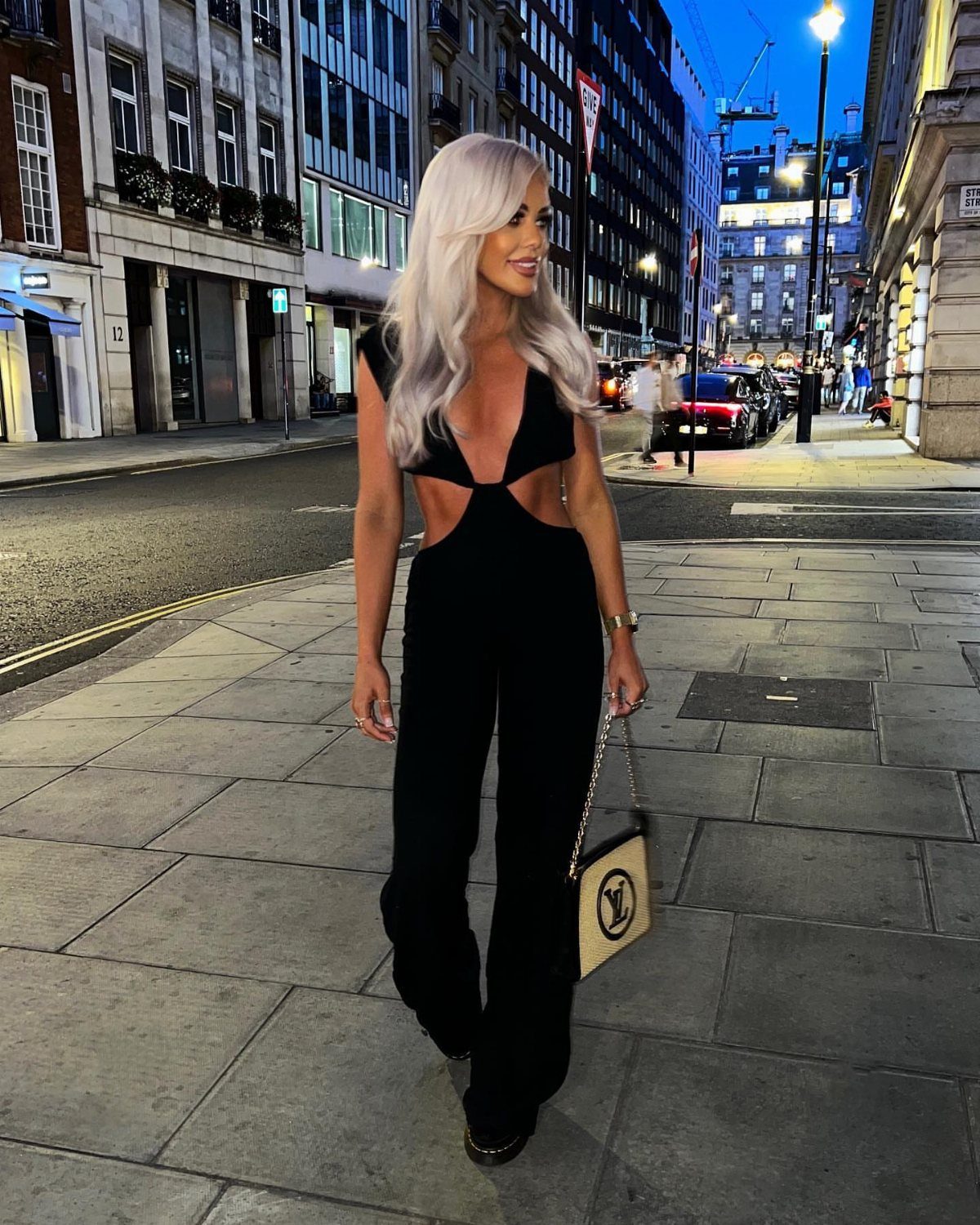 A blonde woman wearing a black cutout jumpsuit standing on a city sidewalk at night.