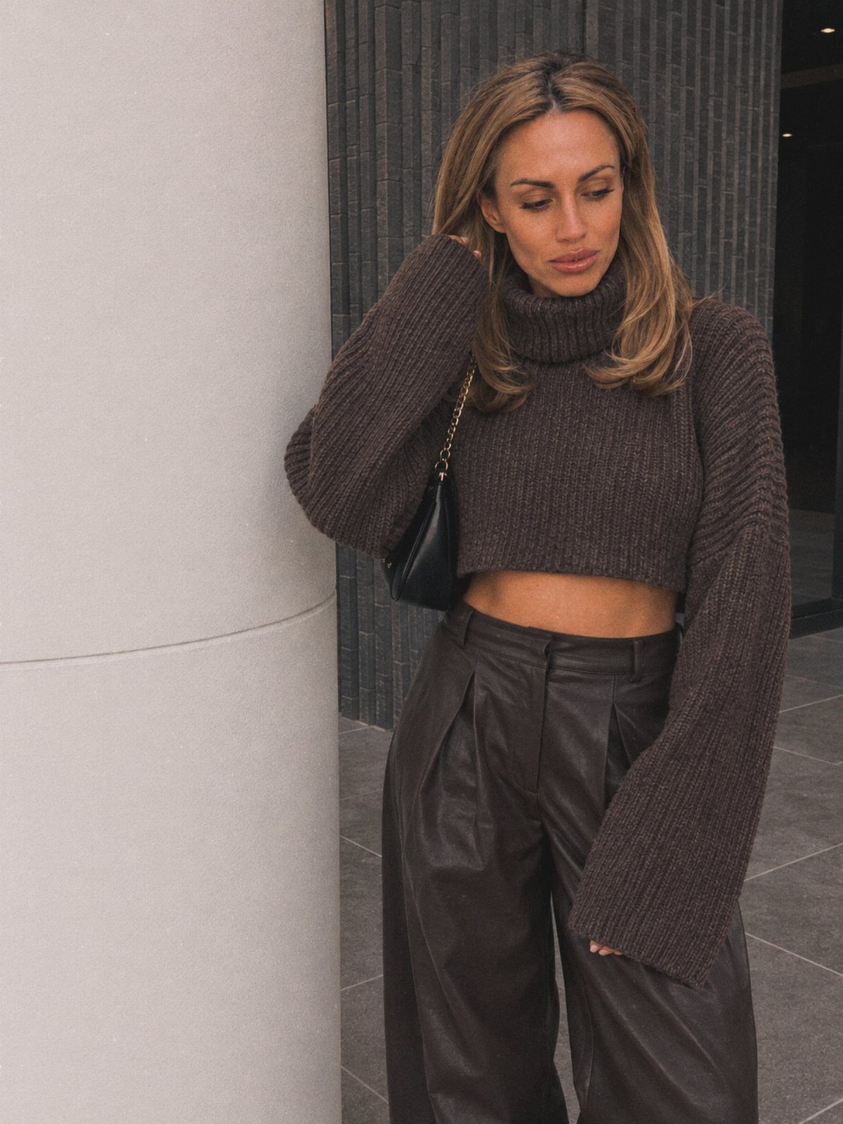 A woman wearing a brown cropped turtleneck sweater and wide-leg leather pants leaning against a pillar.