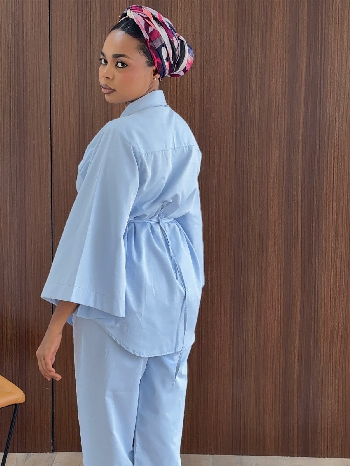 A woman wearing a light blue matching set with a patterned headwrap against wood paneling.