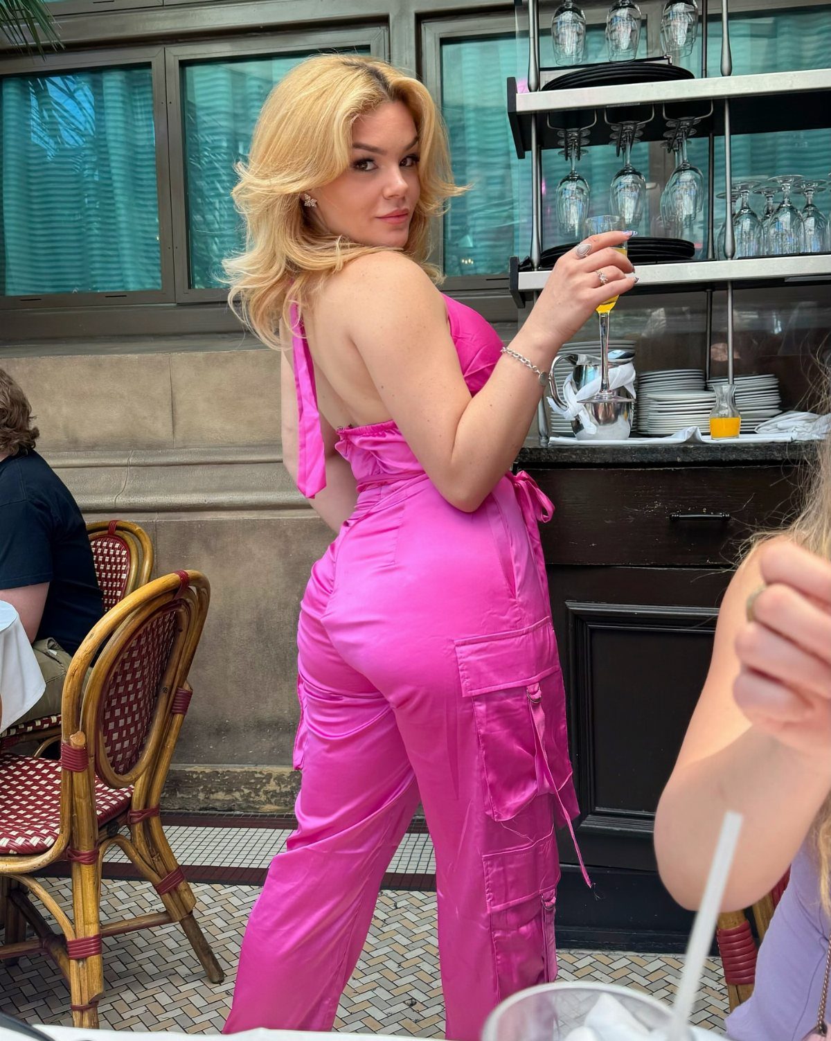 Blonde woman wearing a bright pink satin jumpsuit standing at an outdoor cafe.