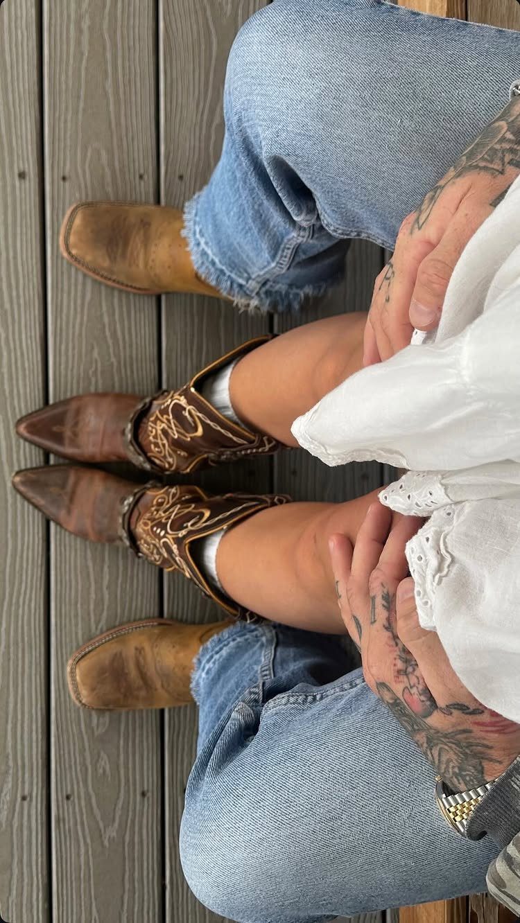 Two people sitting on a wooden deck wearing denim jeans and brown leather cowboy boots.