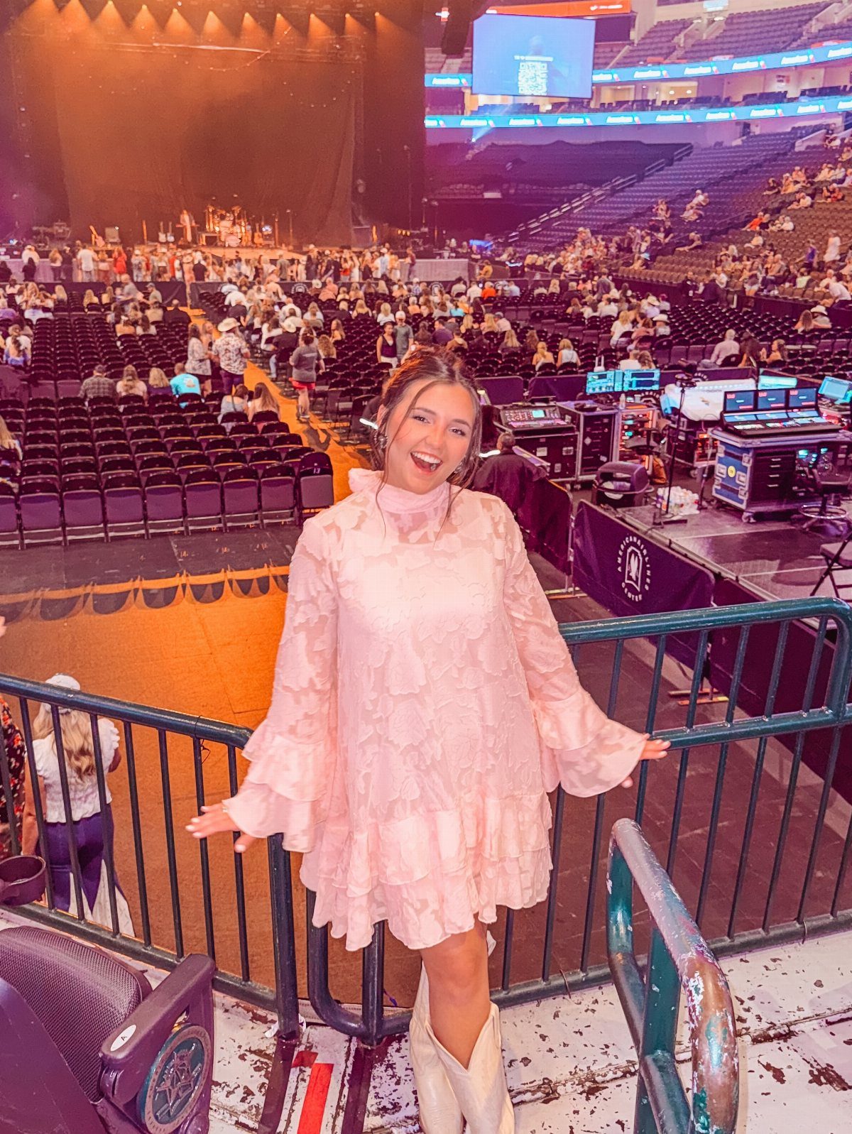A smiling woman in a pink ruffled mini dress and white cowboy boots at a concert arena.