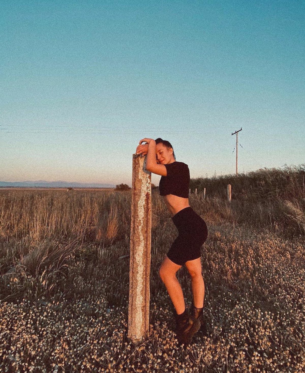 A woman wearing black biker shorts and a crop top leans against a wooden post in a field.