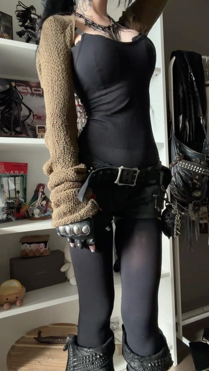 A person wearing a black bodysuit, textured shrug, and chunky black boots standing indoors.