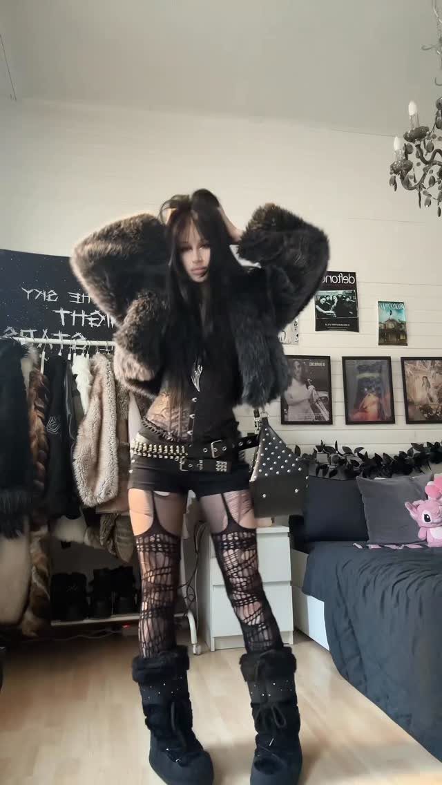 A woman posing in a dark outfit with a large fur coat and fishnet stockings.