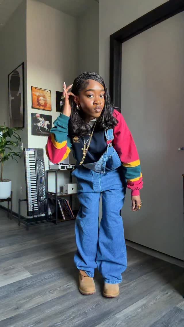 A young woman posing in blue denim overalls and a colorful striped long-sleeved shirt.