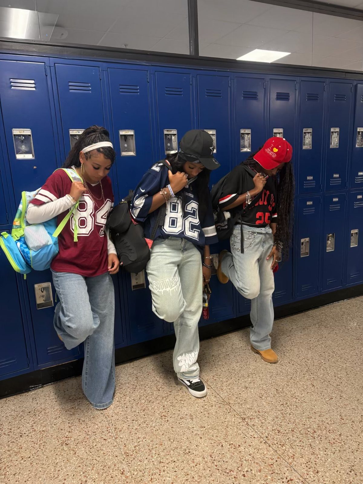 Three young women posing in a school hallway wearing baggy 90s style clothing and jerseys.