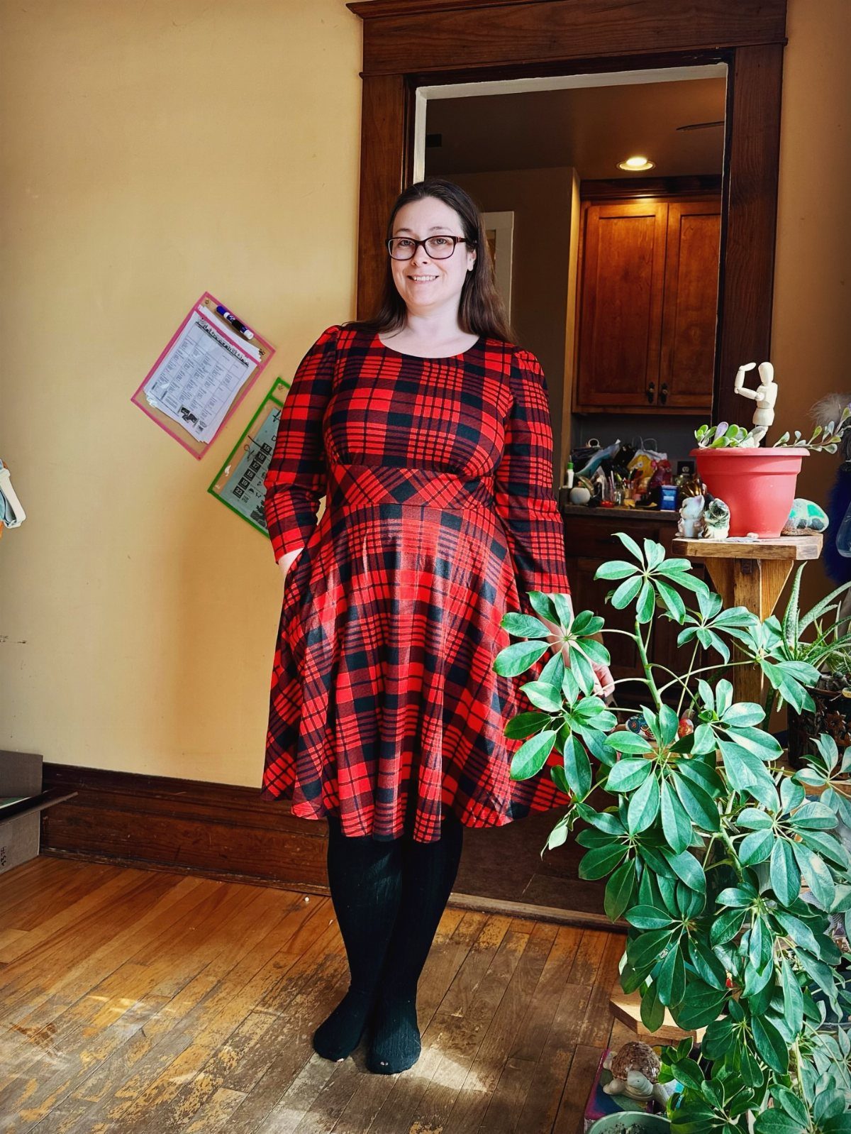 A woman smiling while wearing a red and black plaid dress and black tights indoors.