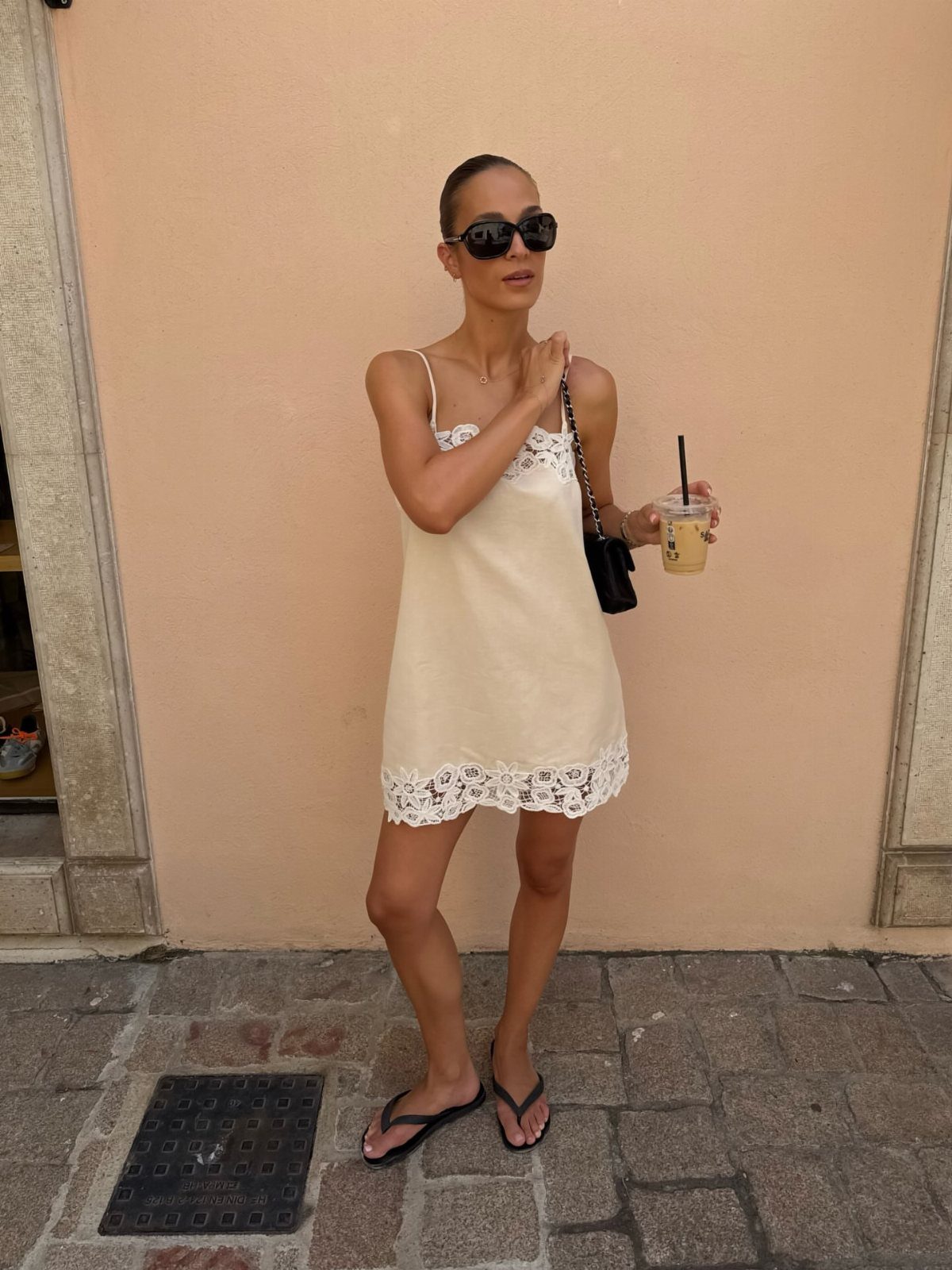 Woman in a white lace slip dress and tiny black sunglasses standing against a peach wall.