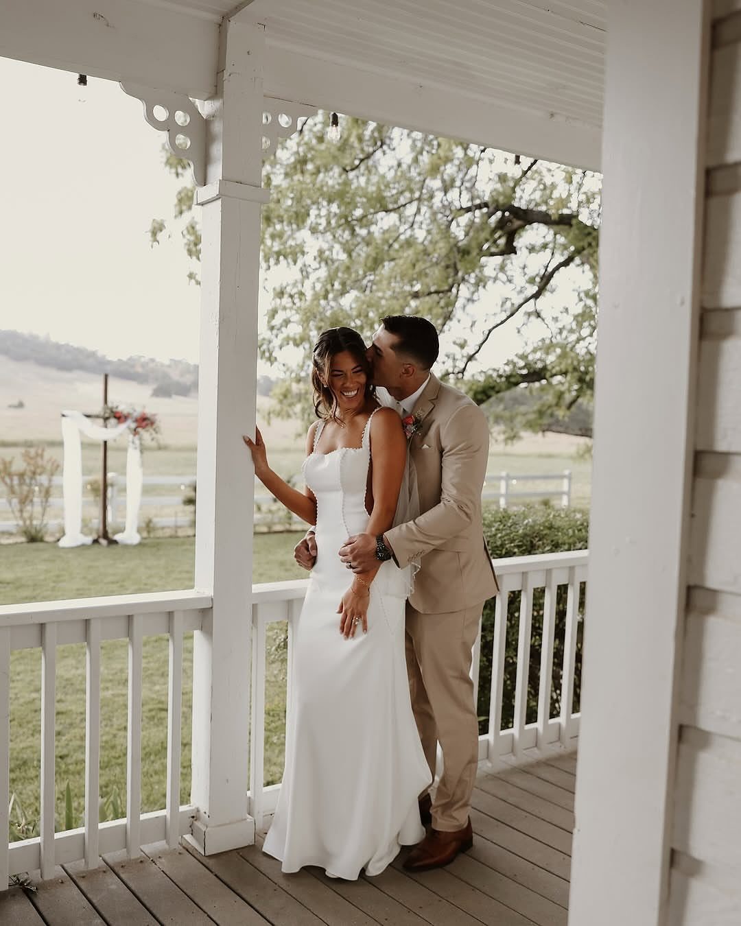 A bride in a white wedding dress and a groom in a tan suit posing on a porch.