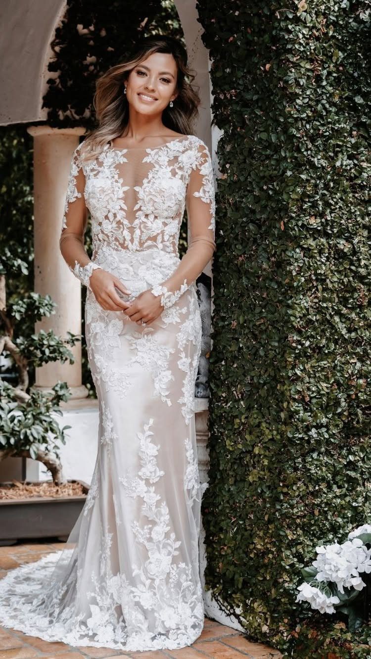 A smiling woman posing in a white lace, long-sleeved sheath wedding dress outdoors.