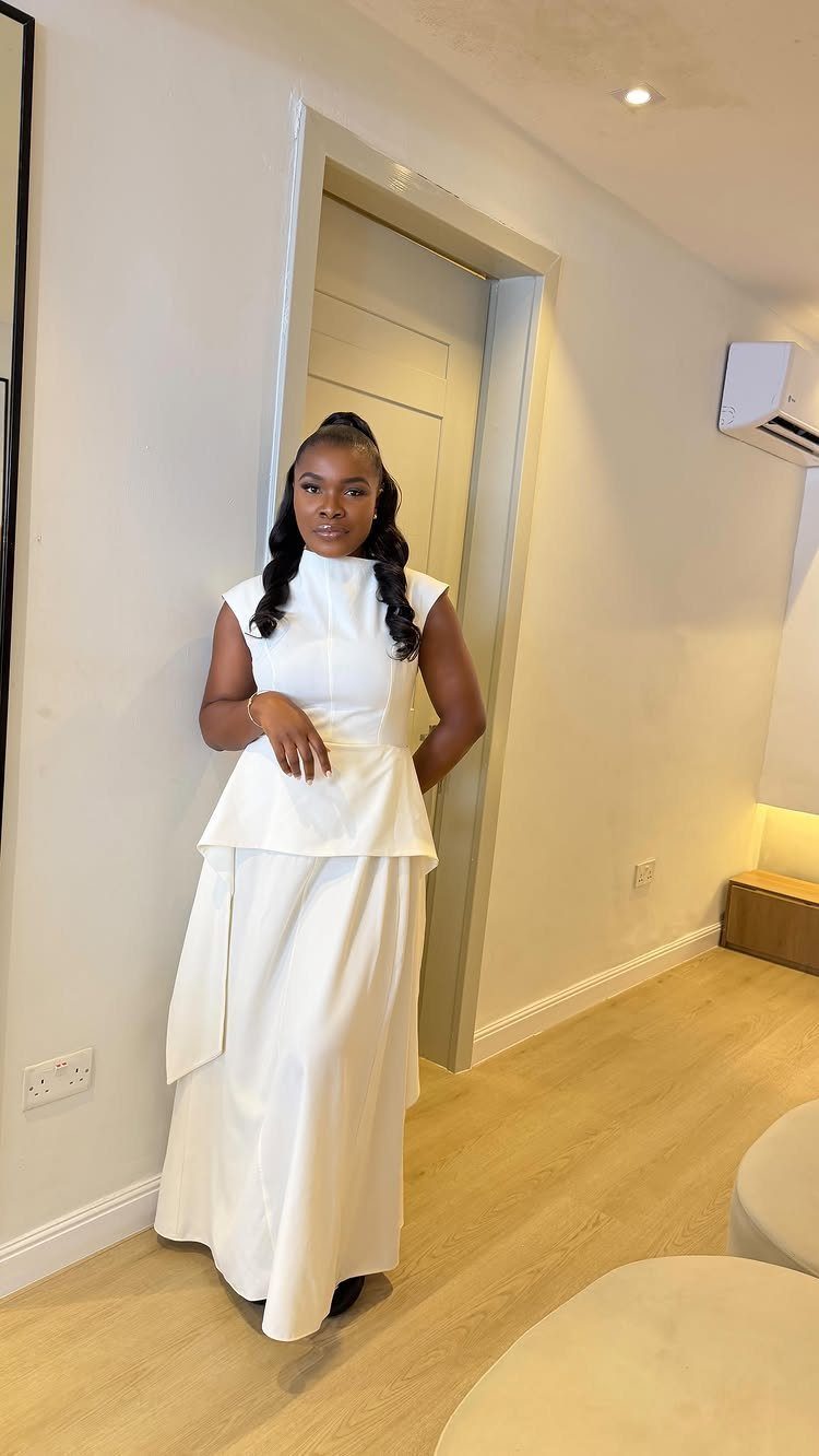 A woman posing in a sleeveless white peplum maxi dress against a neutral indoor background.