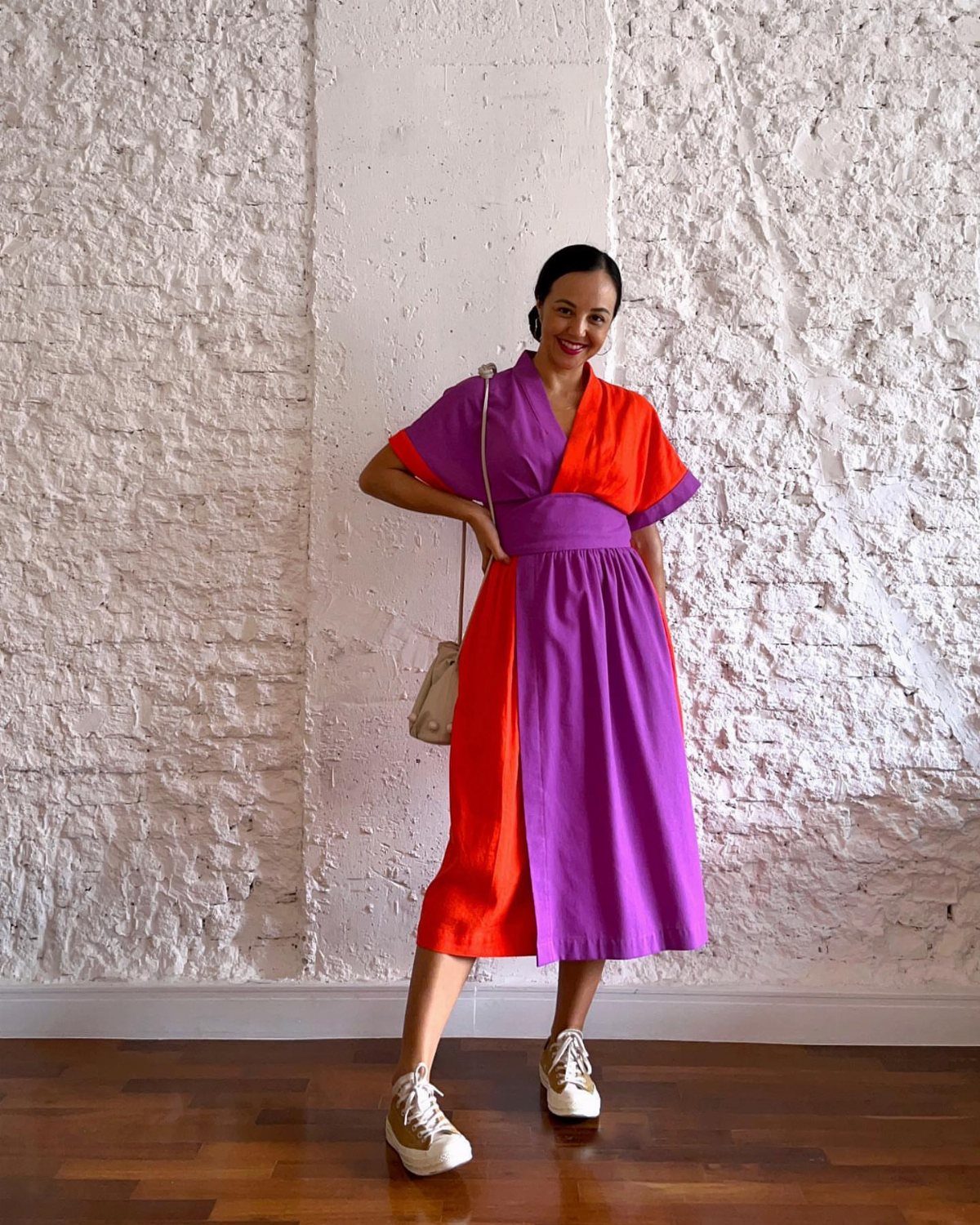 A woman wearing a purple and orange color block wrap dress standing against a white brick wall.