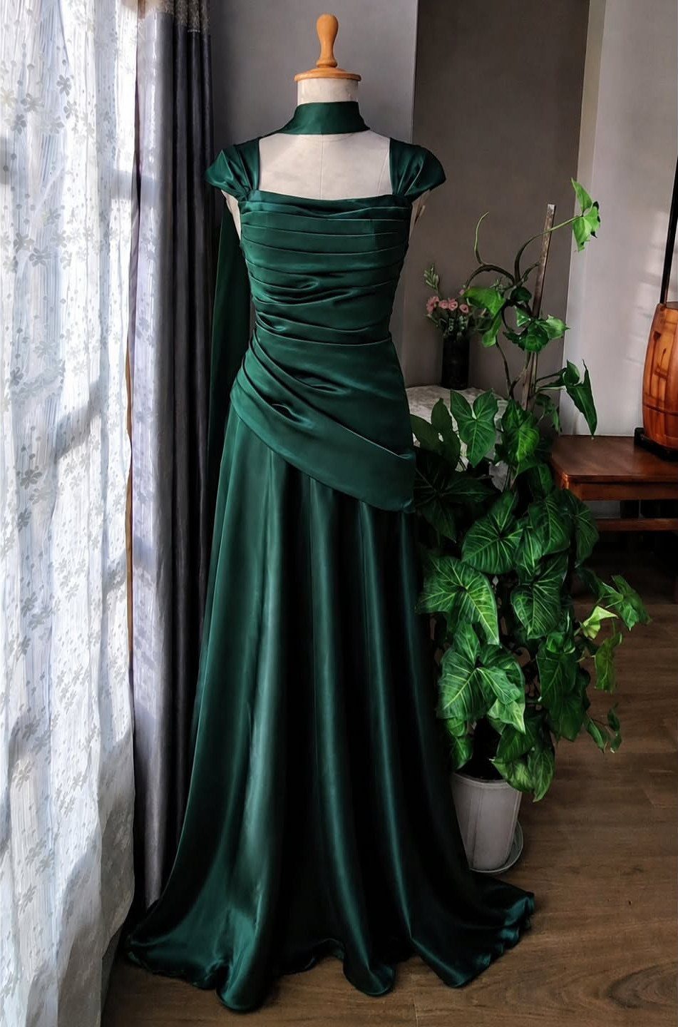 Emerald green satin gown with a ruched bodice displayed on a mannequin near a plant.