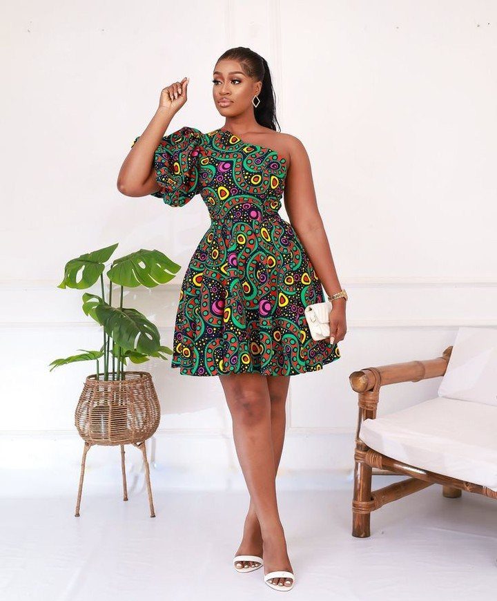 A woman wearing a colorful one-shoulder patterned mini dress standing in a bright room.