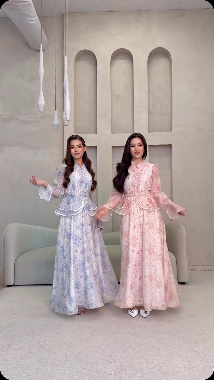 Two women posing in long, floral patterned dresses with ruffled waistlines in a minimalist room.