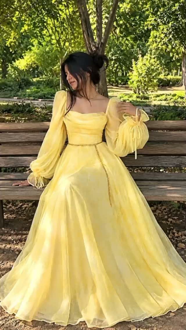A woman in a long yellow puff-sleeved dress sitting on a wooden park bench.
