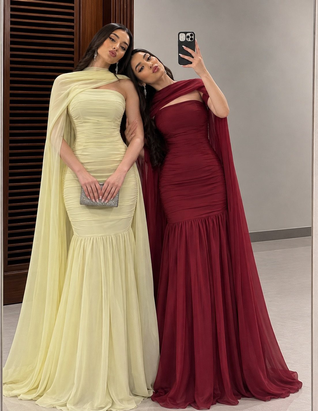 Two women posing in long evening gowns with draped fabric wraps over their shoulders.