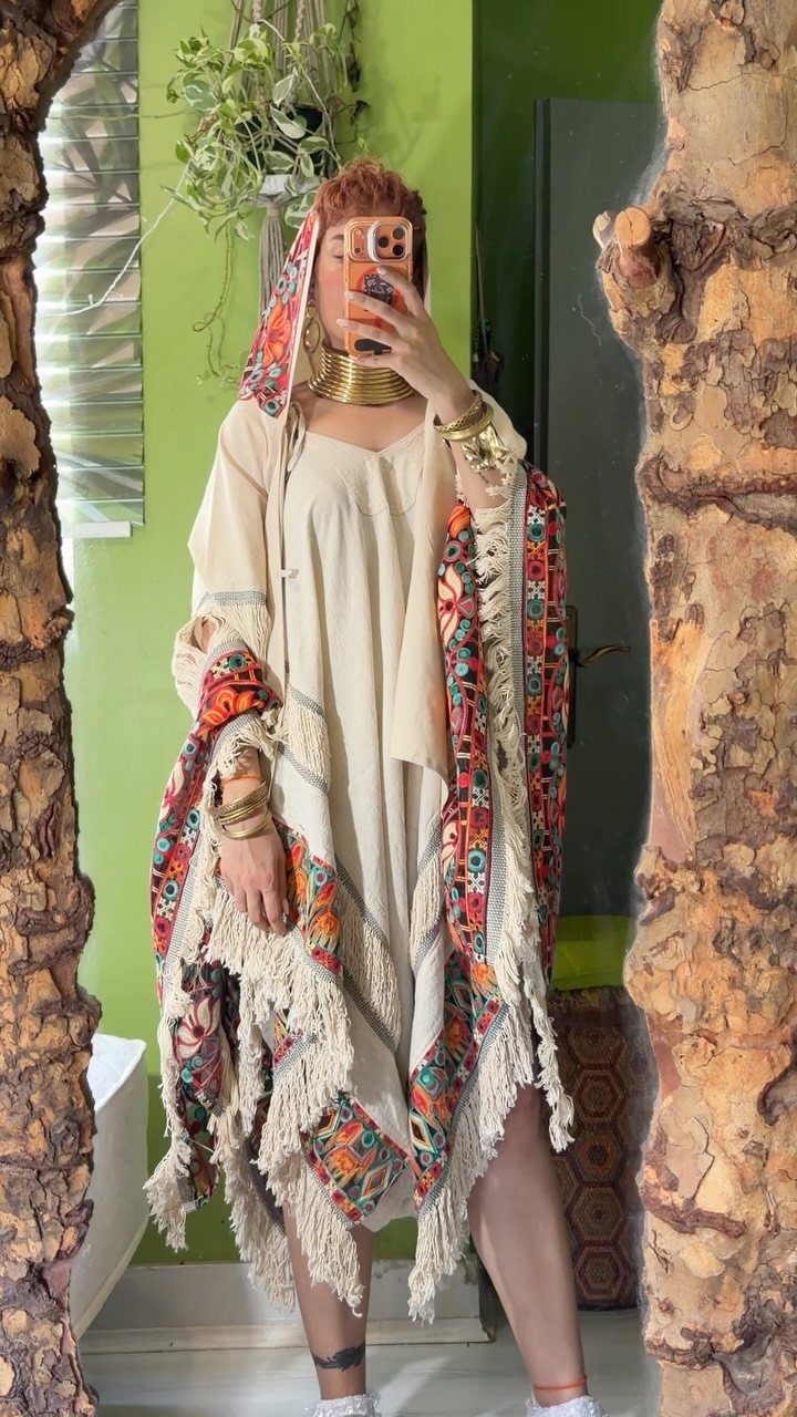 A person wearing a cream dress and a colorful fringed bohemian shawl posing for a selfie.