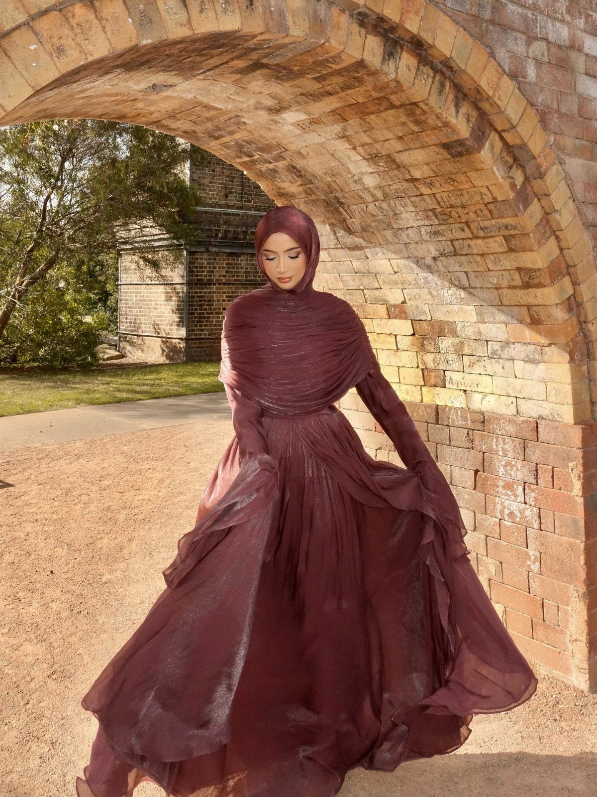 A woman in a flowing mauve dress and matching shawl standing under a brick archway.
