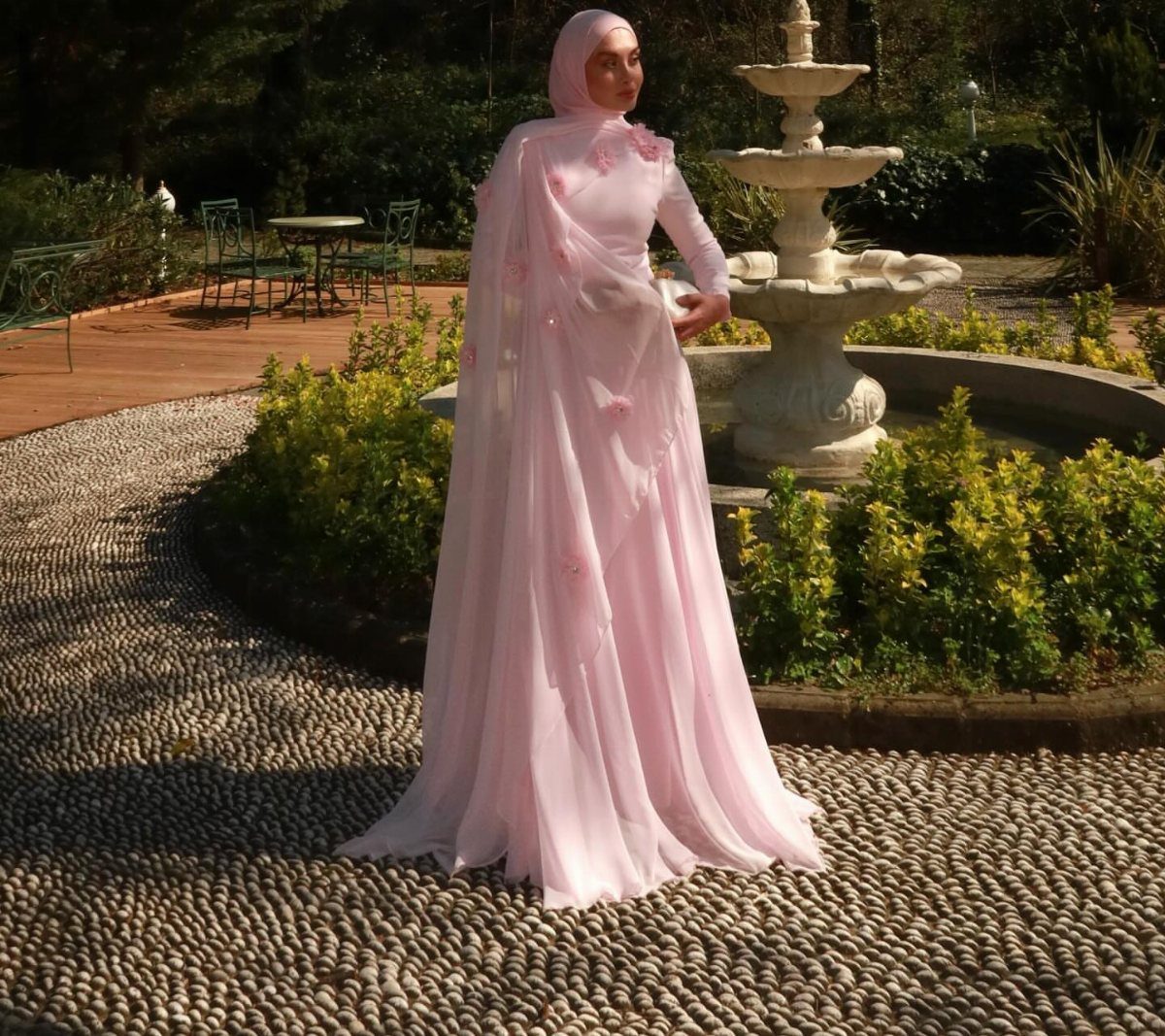 A woman in a long pink dress and matching floral shawl standing by a fountain.