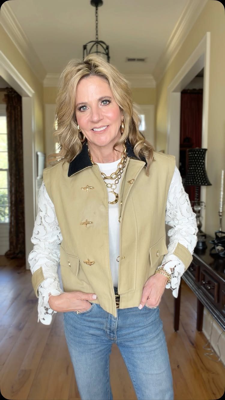 A smiling blonde woman wearing a tan vest, white lace shirt, and blue jeans.