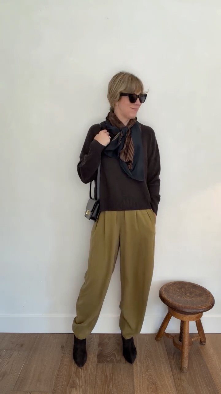 A woman in olive trousers and a dark sweater wearing sunglasses and a patterned scarf.