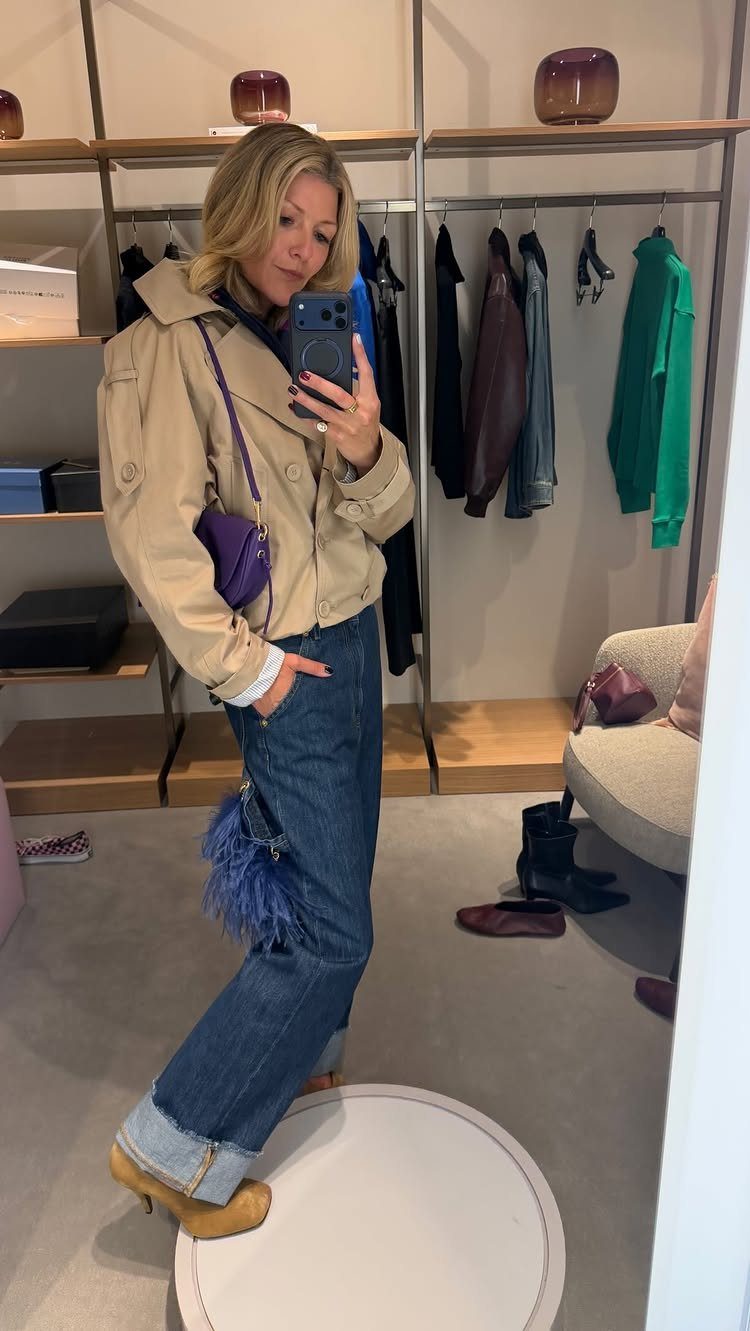 A woman in a tan jacket and blue jeans posing for a mirror selfie in a boutique.