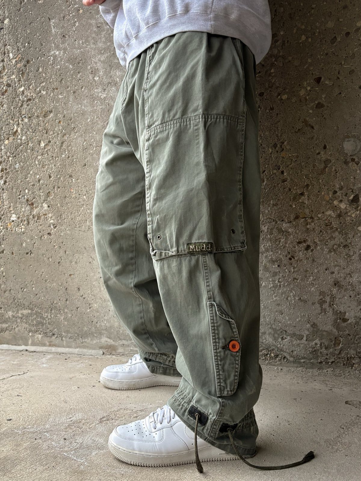 Olive green baggy cargo pants with large pockets paired with white sneakers against a concrete wall.